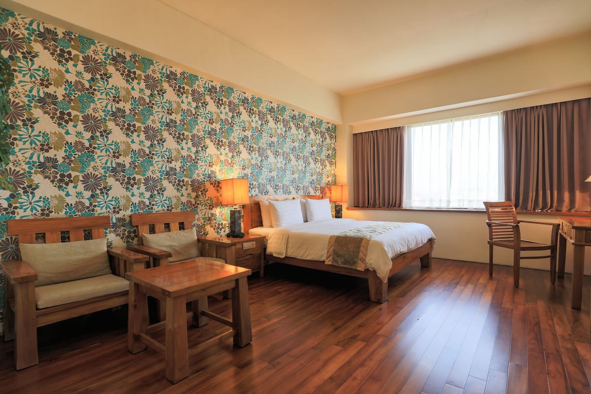 A hotel room with a wooden floor, a bed with white linens, a wooden bench, a small table, and a wall with a floral pattern.