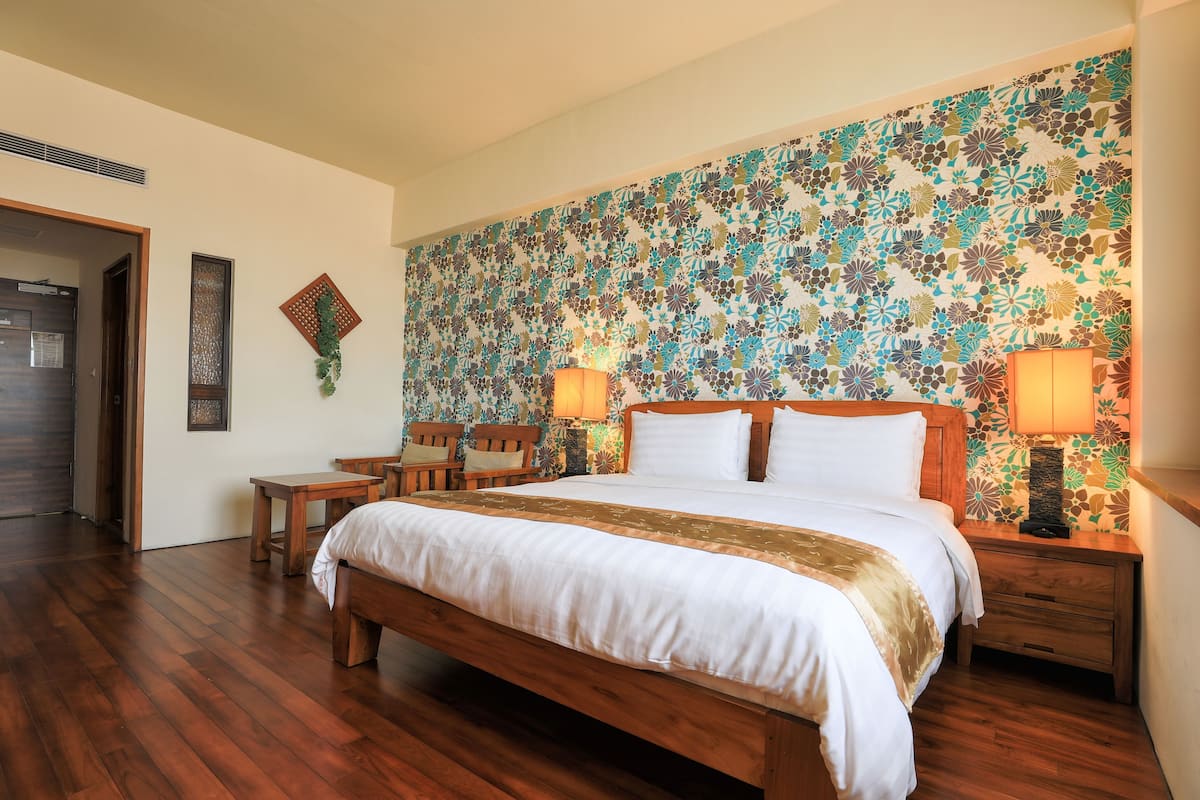 A hotel room with a wooden bed, two bedside tables, a nightstand, a patterned wall, and a door leading to another room.