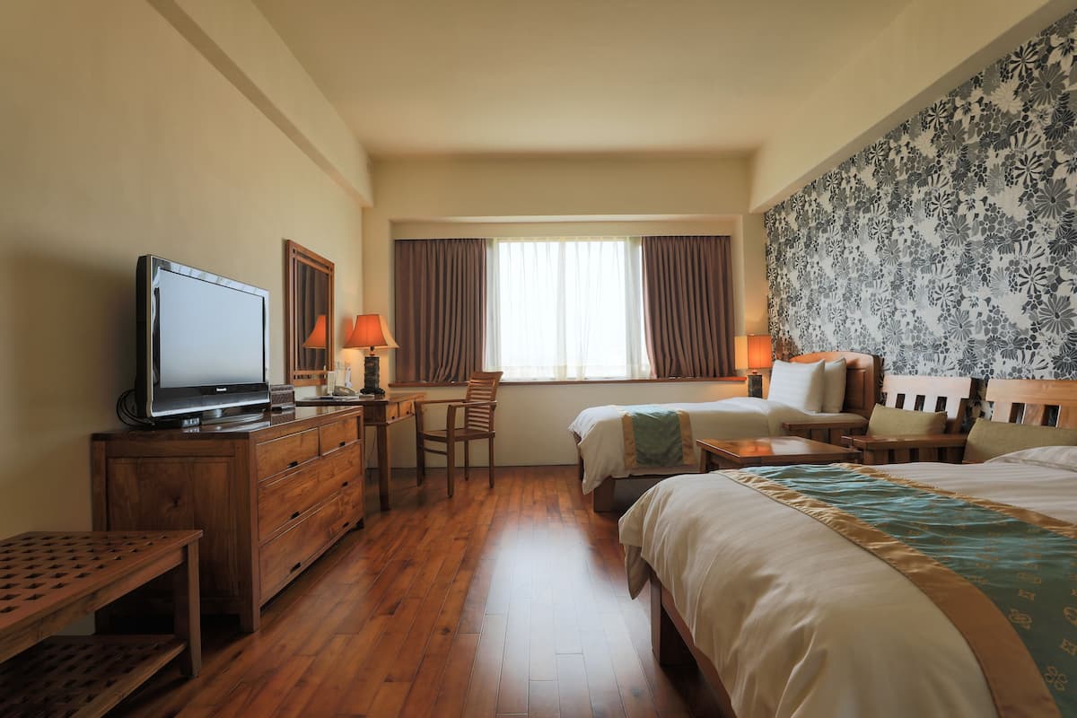 A hotel room with a wooden floor, a TV, a desk with a lamp, two beds, and a large floral-patterned wall.