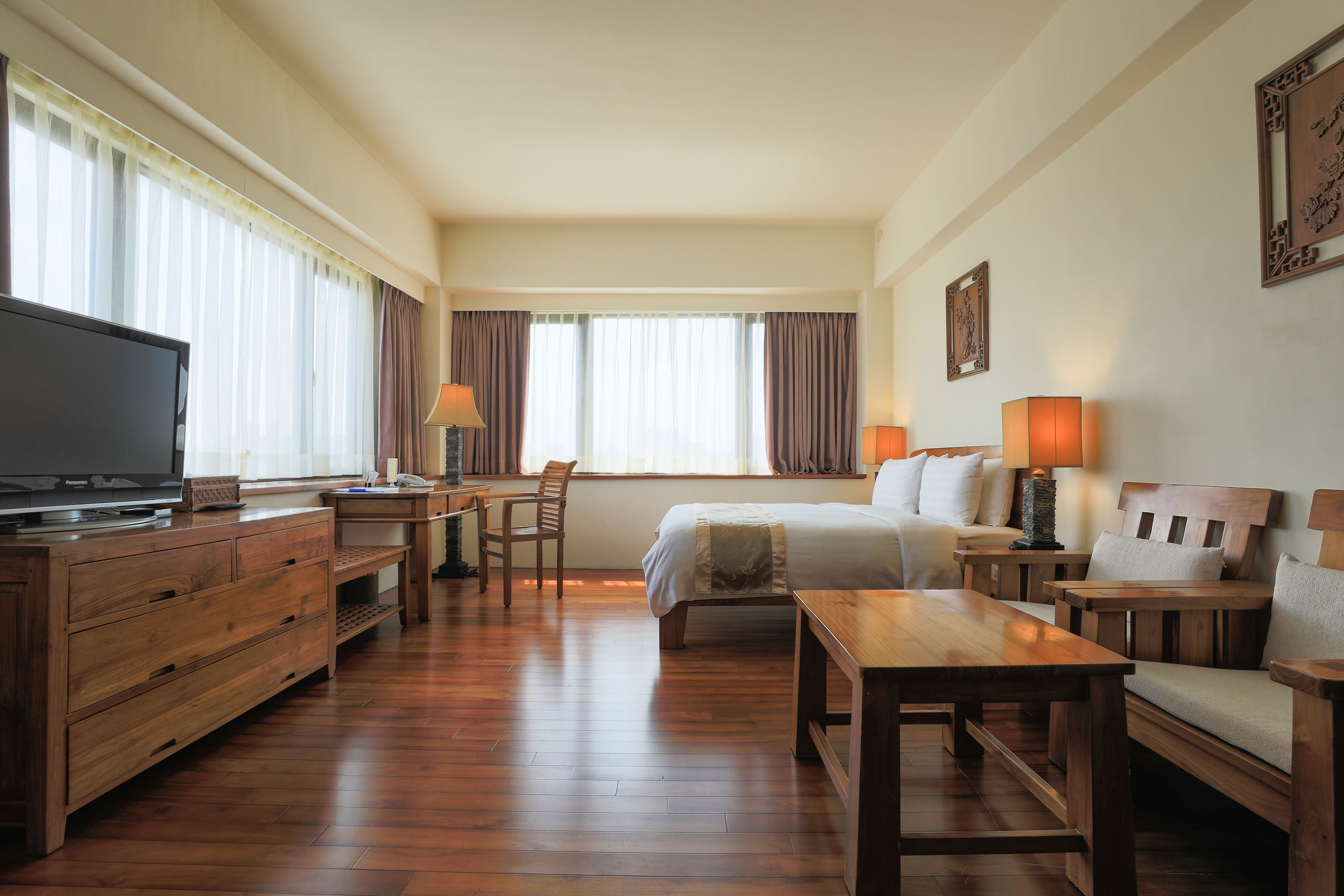 A hotel room with a wooden floor, a bed, a desk, a chair, a TV, and two lamps.
