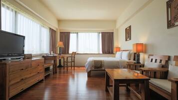 A hotel room with a wooden floor, a bed, a desk, a chair, a TV, and two lamps.
