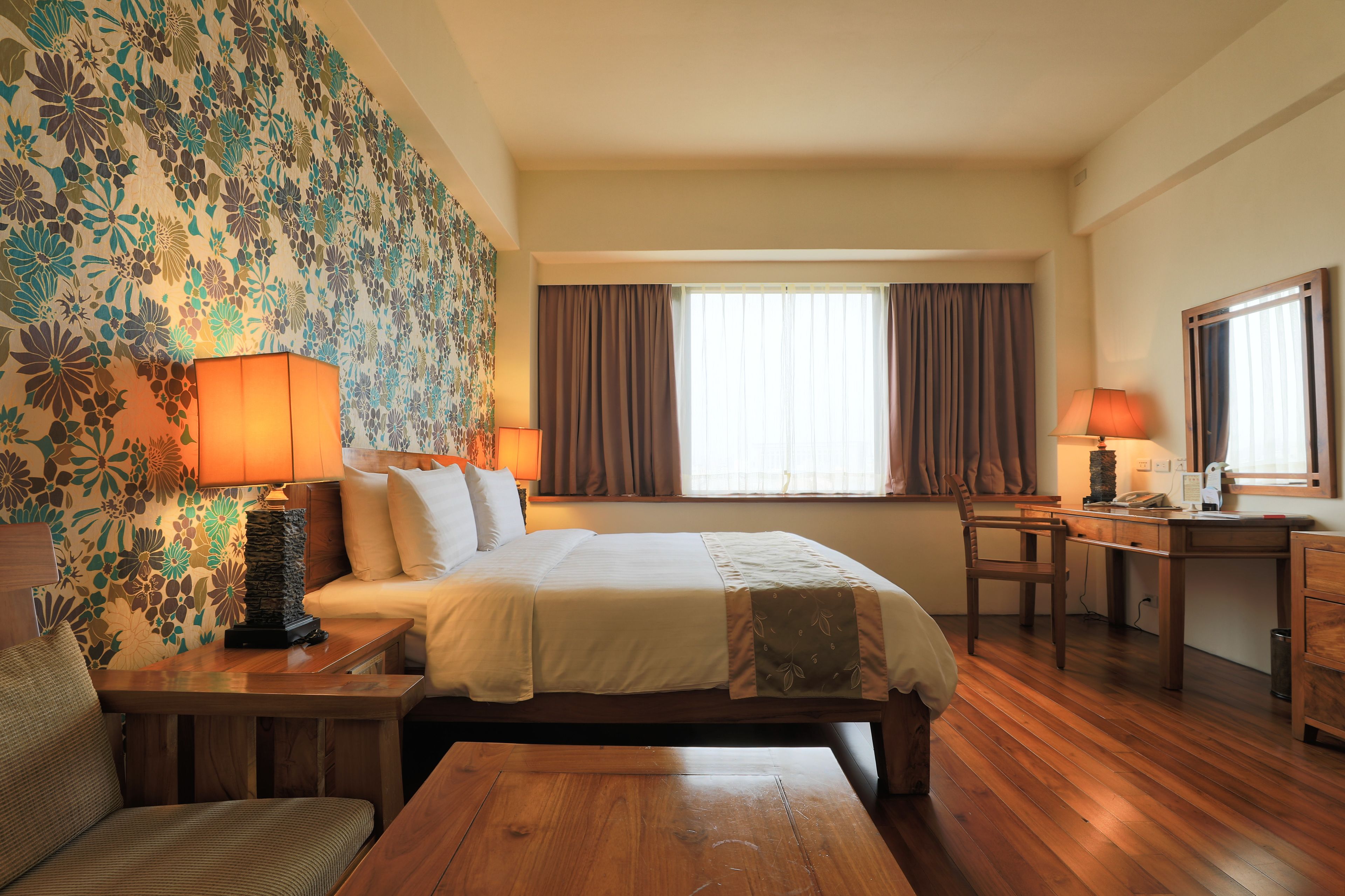 Una habitación de hotel con cama, escritorio, silla, lámpara y ventana con cortinas.