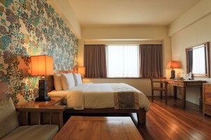 Una habitación de hotel con cama, escritorio, silla, lámpara y ventana con cortinas.