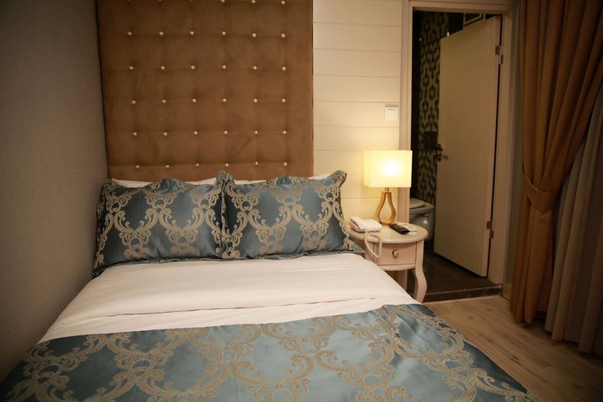 A hotel room with a tufted headboard, a bed with patterned bedding, a bedside table with a lamp, and a door with curtains.