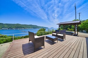 A wooden deck with wicker furniture and a gazebo overlooking a coastal landscape with hills and a body of water. - Tsubaki House (Anan)