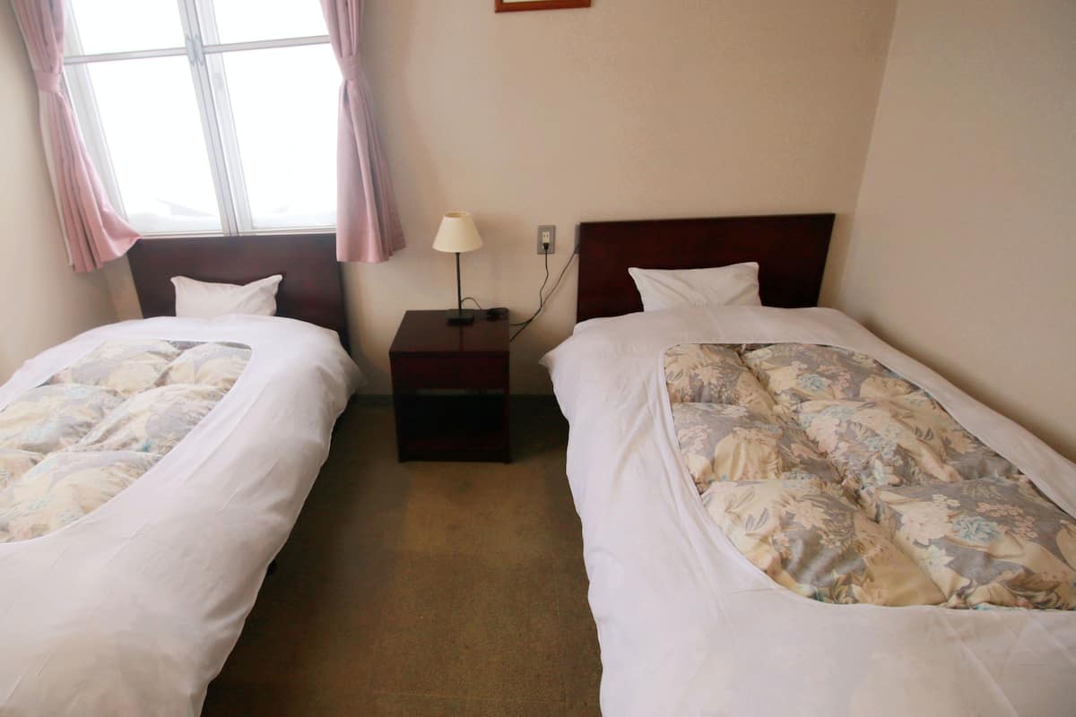 A hotel room with two single beds, a nightstand with a lamp, and a window with curtains.
