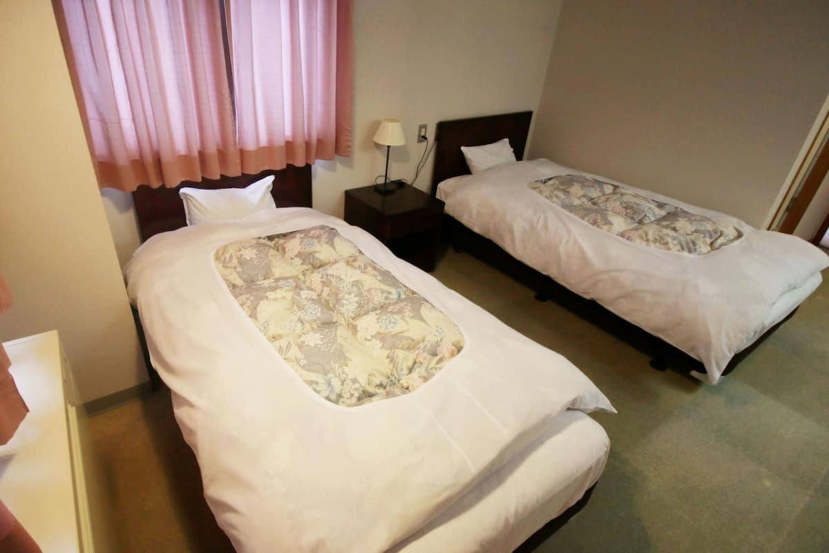 A hotel room with two single beds, a nightstand with a lamp, and a window with pink curtains.