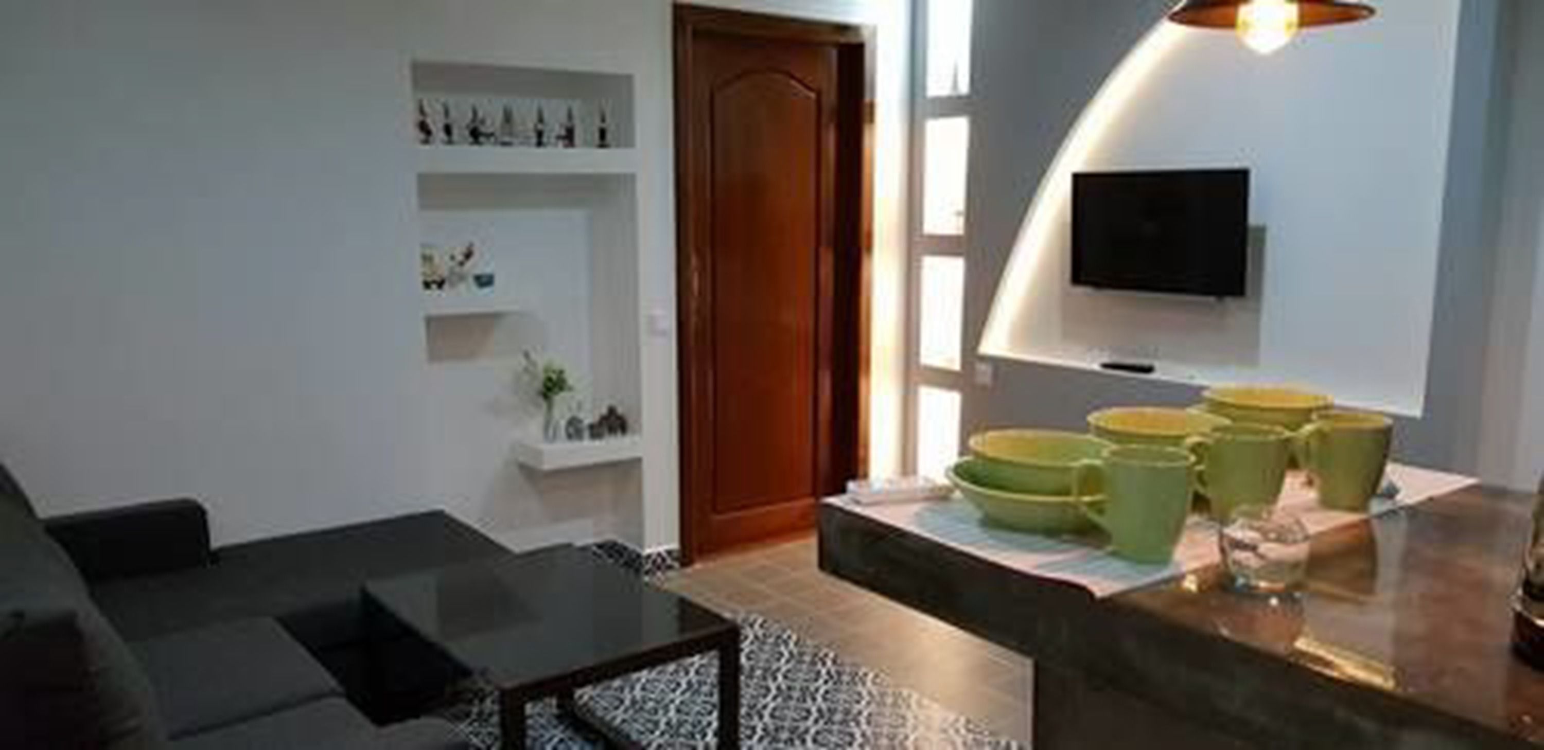 A modern living room with a sofa, a coffee table, and a TV mounted on the wall. There are shelves with decorative items and a door leading to another room.