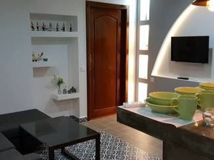 Un salon moderne avec un canapé, une table basse et un téléviseur fixé au mur. On y trouve des étagères ornées d’objets décoratifs et une porte qui donne sur une autre pièce.