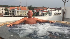 A man in a hot tub surrounded by pebbles, with buildings in the background. - Katy Resident and Spa (Siem Reap)
