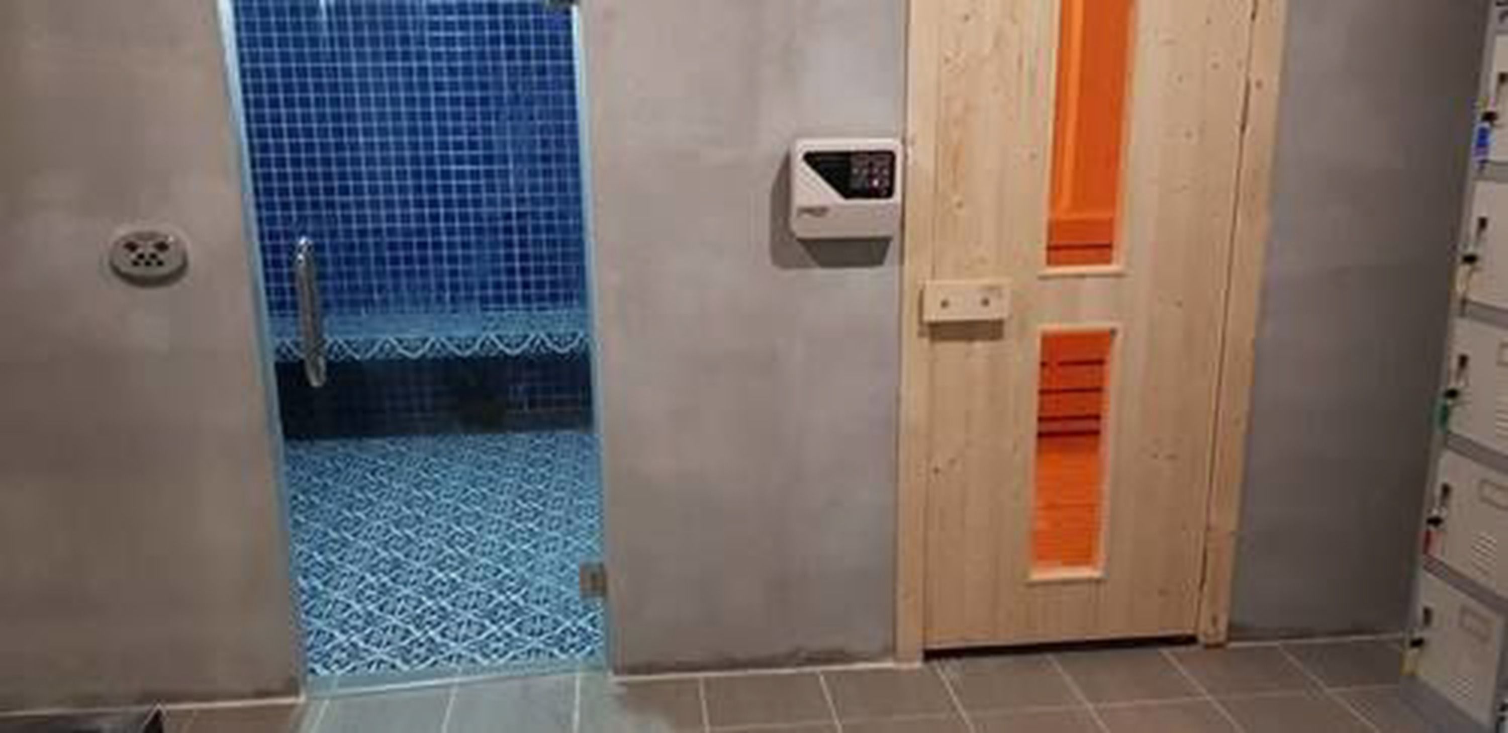 A bathroom with a shower, a toilet, and a sink. There is a wooden door with an orange stripe and a small control panel on the wall.