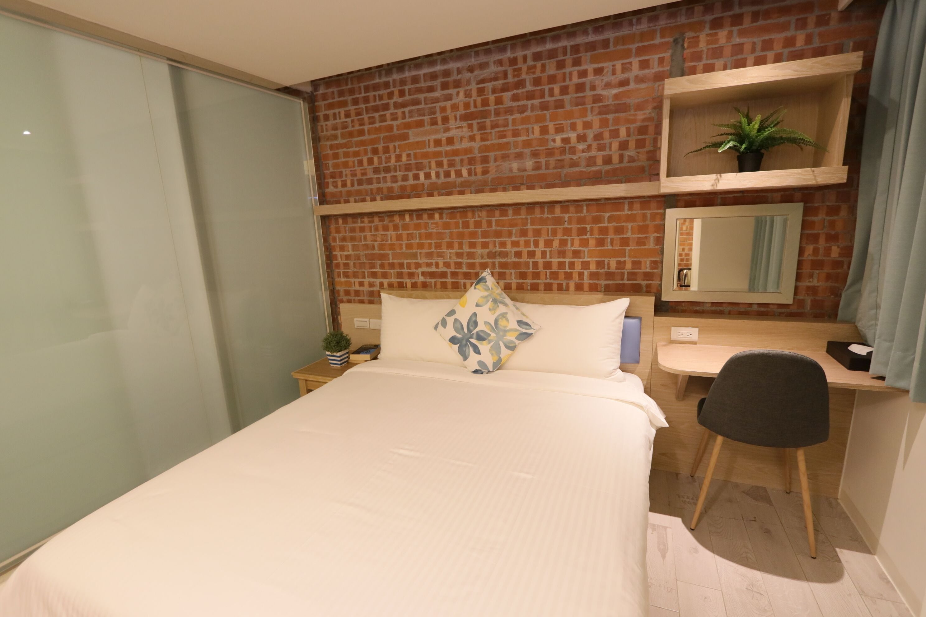 A hotel room with a bed, a desk, a chair, a mirror, and a potted plant.