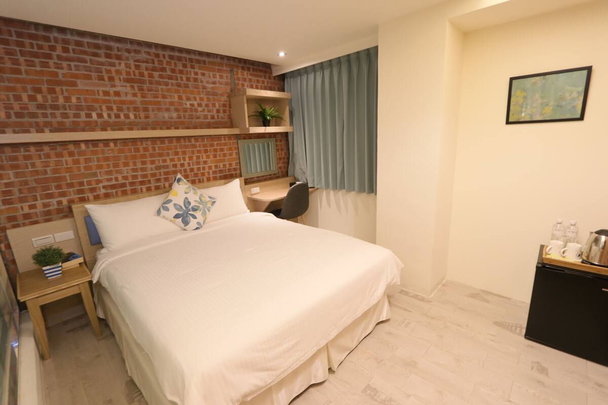 A hotel room with a brick wall, a bed with white bedding, a desk with a chair, a small table with a plant, and a refrigerator.