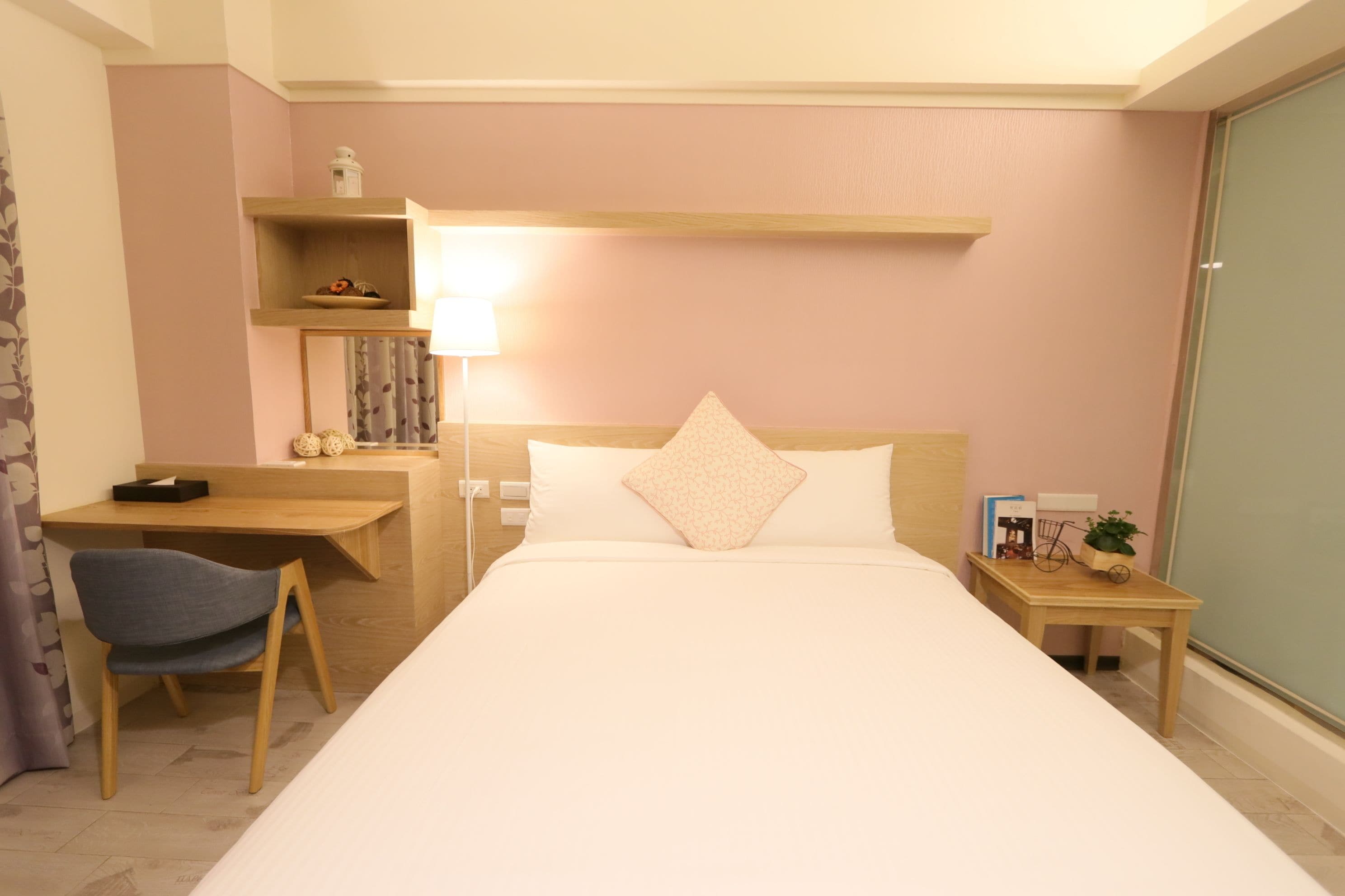 A hotel room with a bed, a desk, a chair, a bedside table, and a lamp.