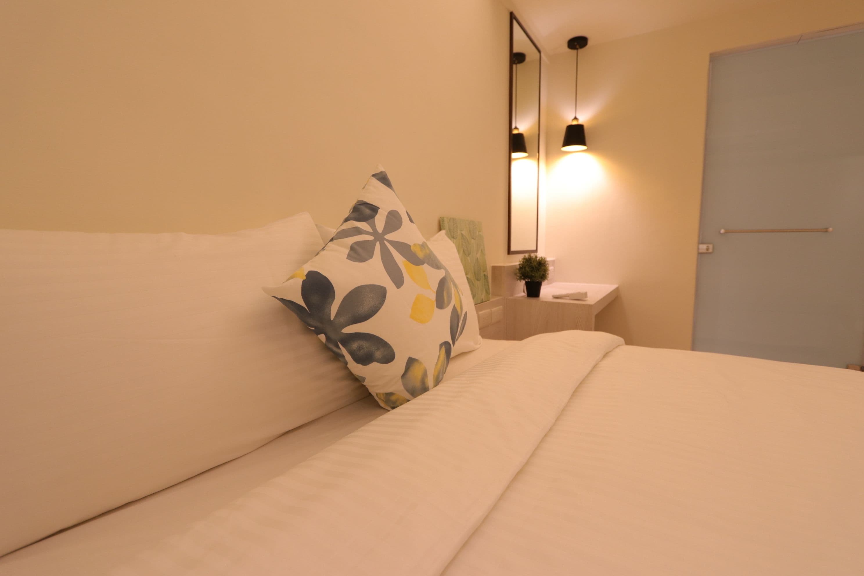 A neatly made bed with a floral pillow, a bedside table with a lamp, and a door in the background.