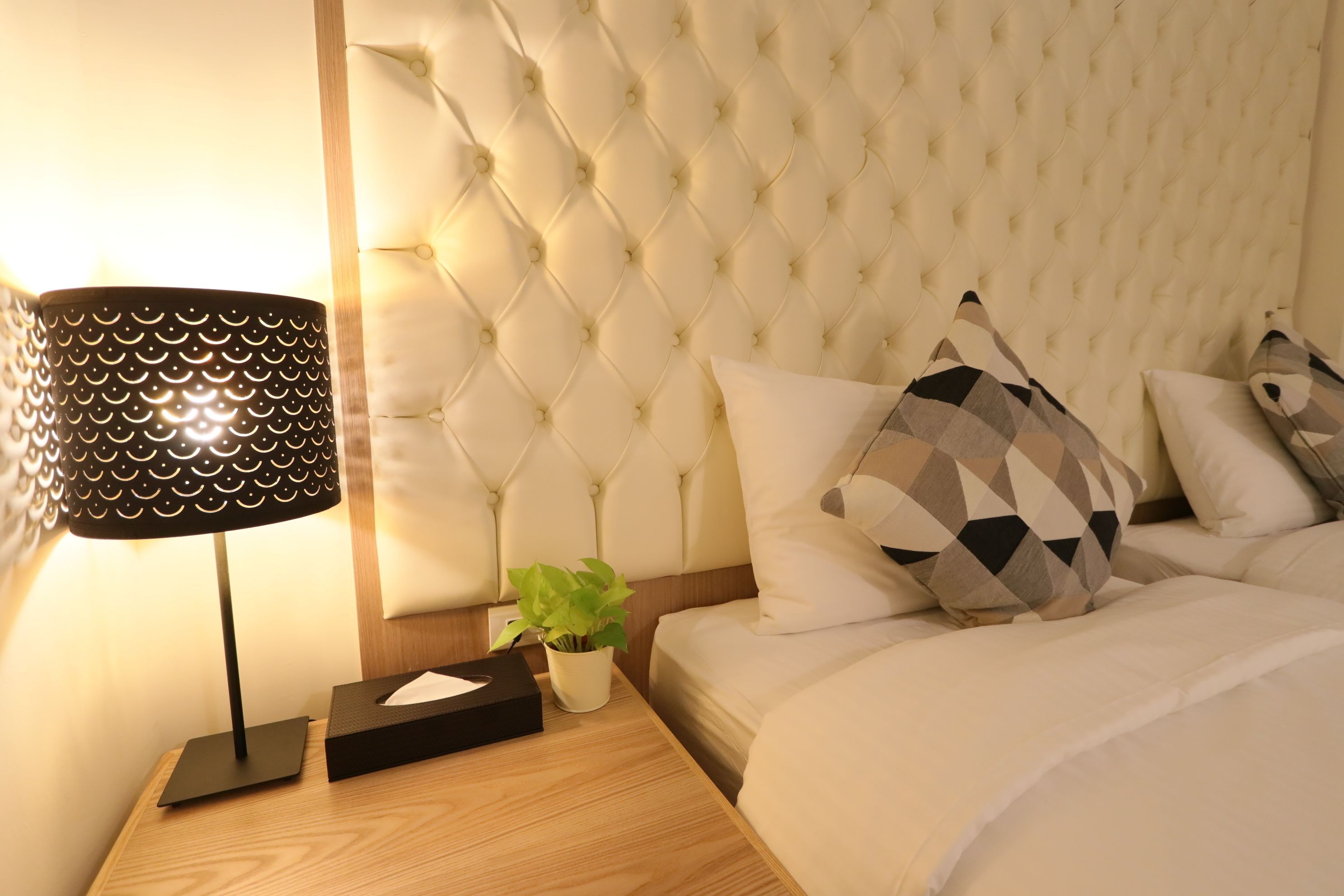 Cama bem arrumada com cabeceira estofada, luminária de cabeceira e uma pequena planta em vaso sobre mesa de cabeceira de madeira.