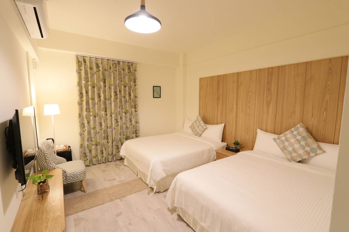 A hotel room with two beds, a wooden headboard, a desk with a lamp, a chair, a TV, and a window with curtains.