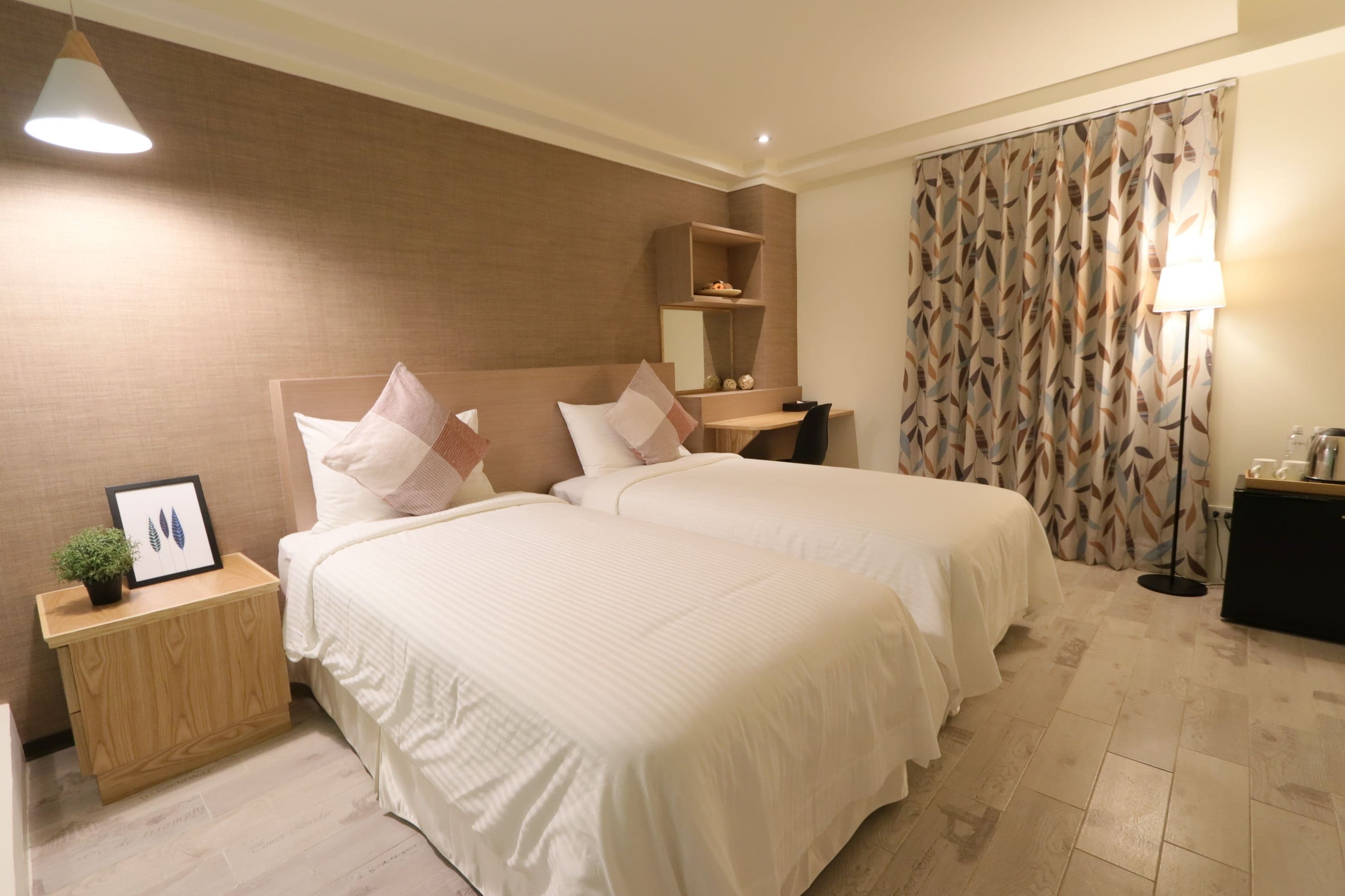 Une chambre d’hôtel avec deux lits, une table de chevet avec une lampe, une petite étagère et une grande fenêtre avec des rideaux.