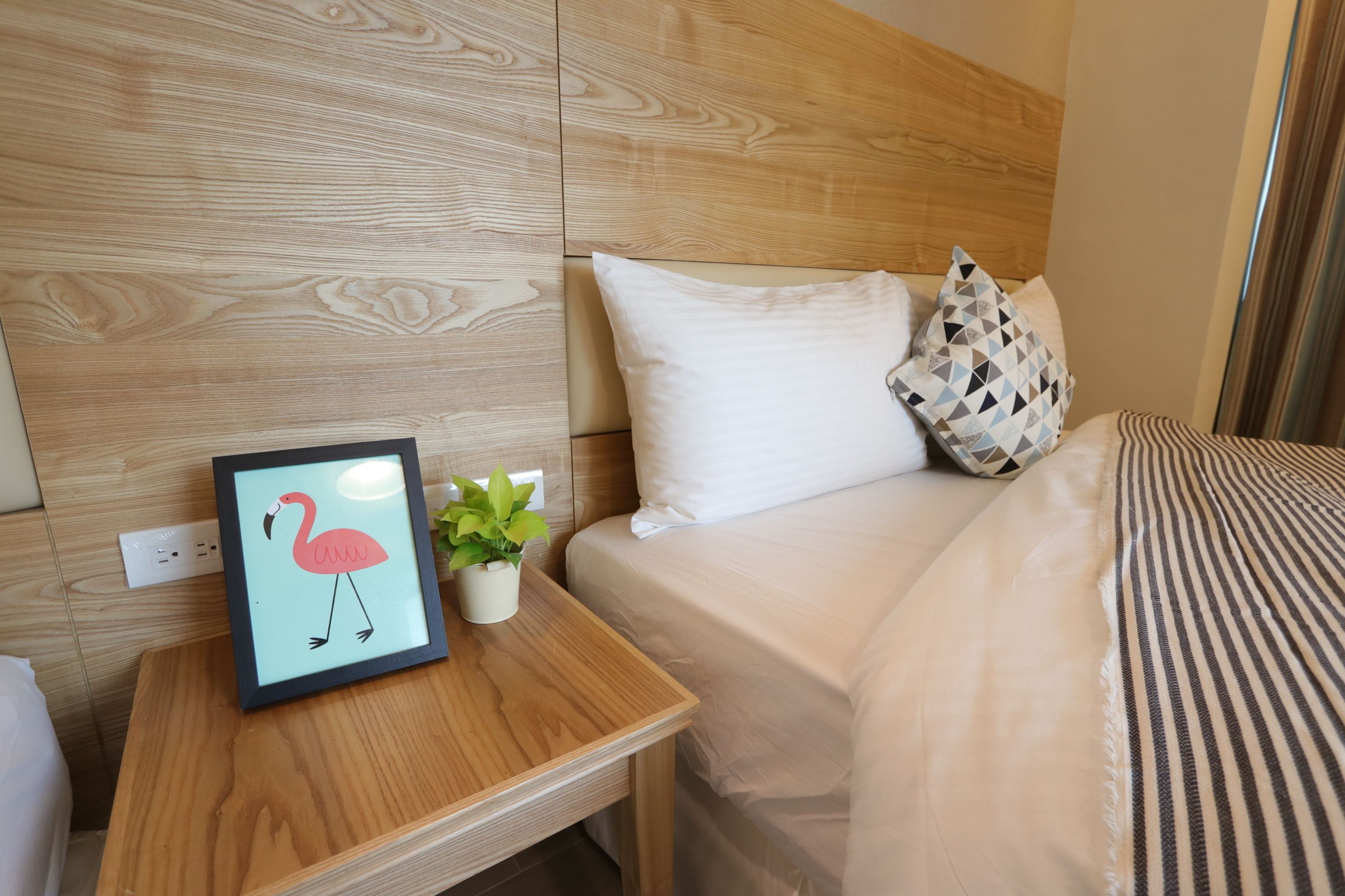 Habitación de hotel con cabecera de madera, cama con ropa de cama blanca, mesita de noche con un cuadro enmarcado de un flamenco y una pequeña planta en maceta.