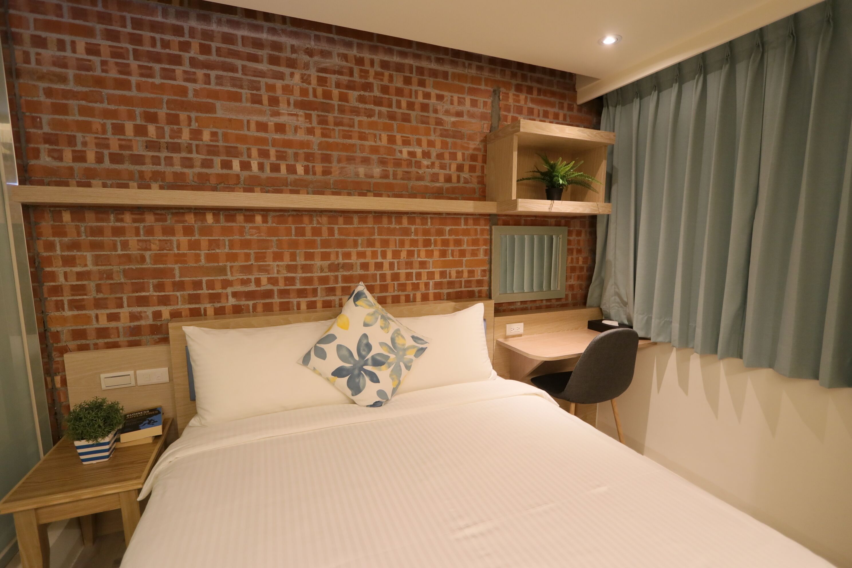 A hotel room with a brick wall, a bed with white bedding, a desk with a chair, and a window with curtains.