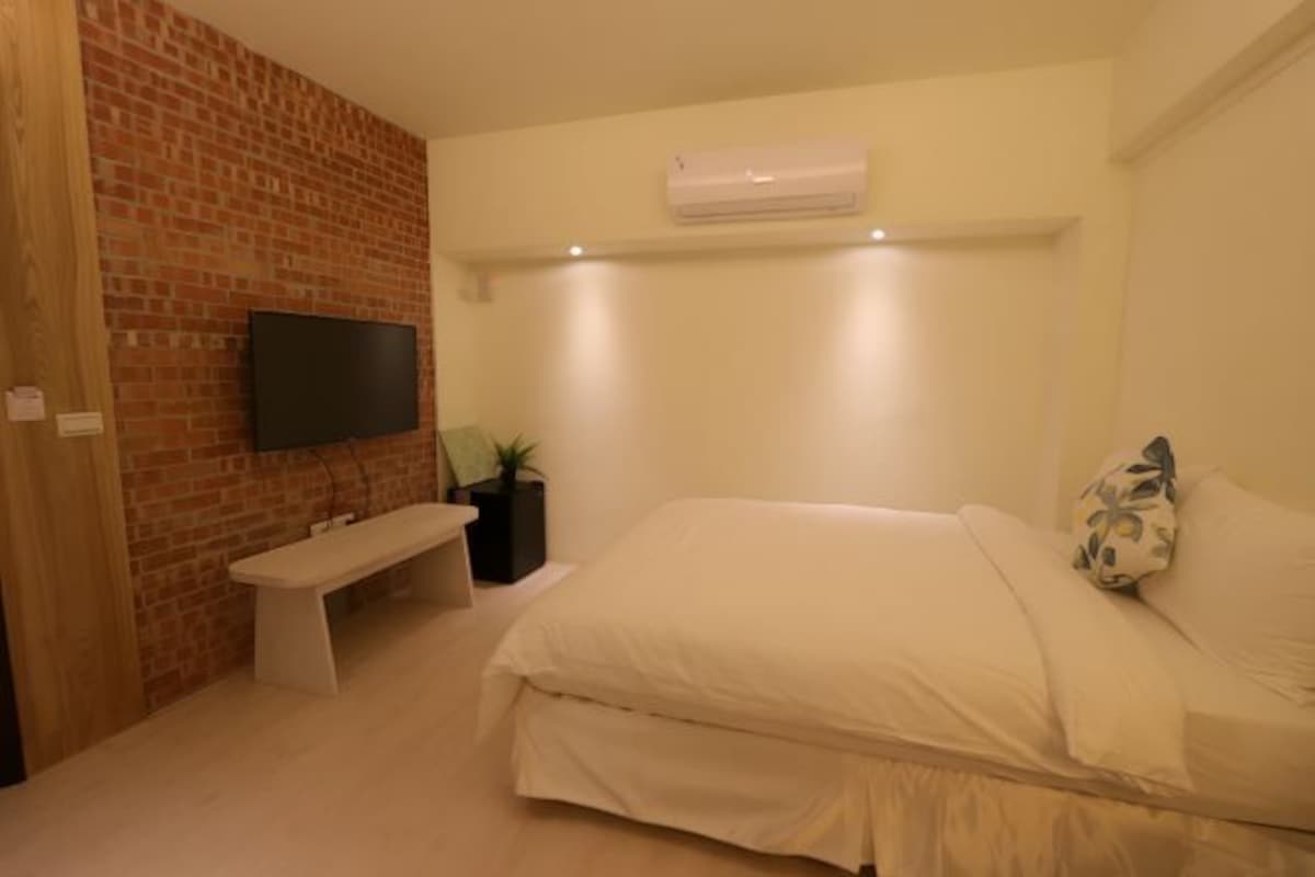 A bedroom with a brick accent wall, a flat-screen TV mounted above a bench, a bed with white bedding, and an air conditioning unit on the wall.