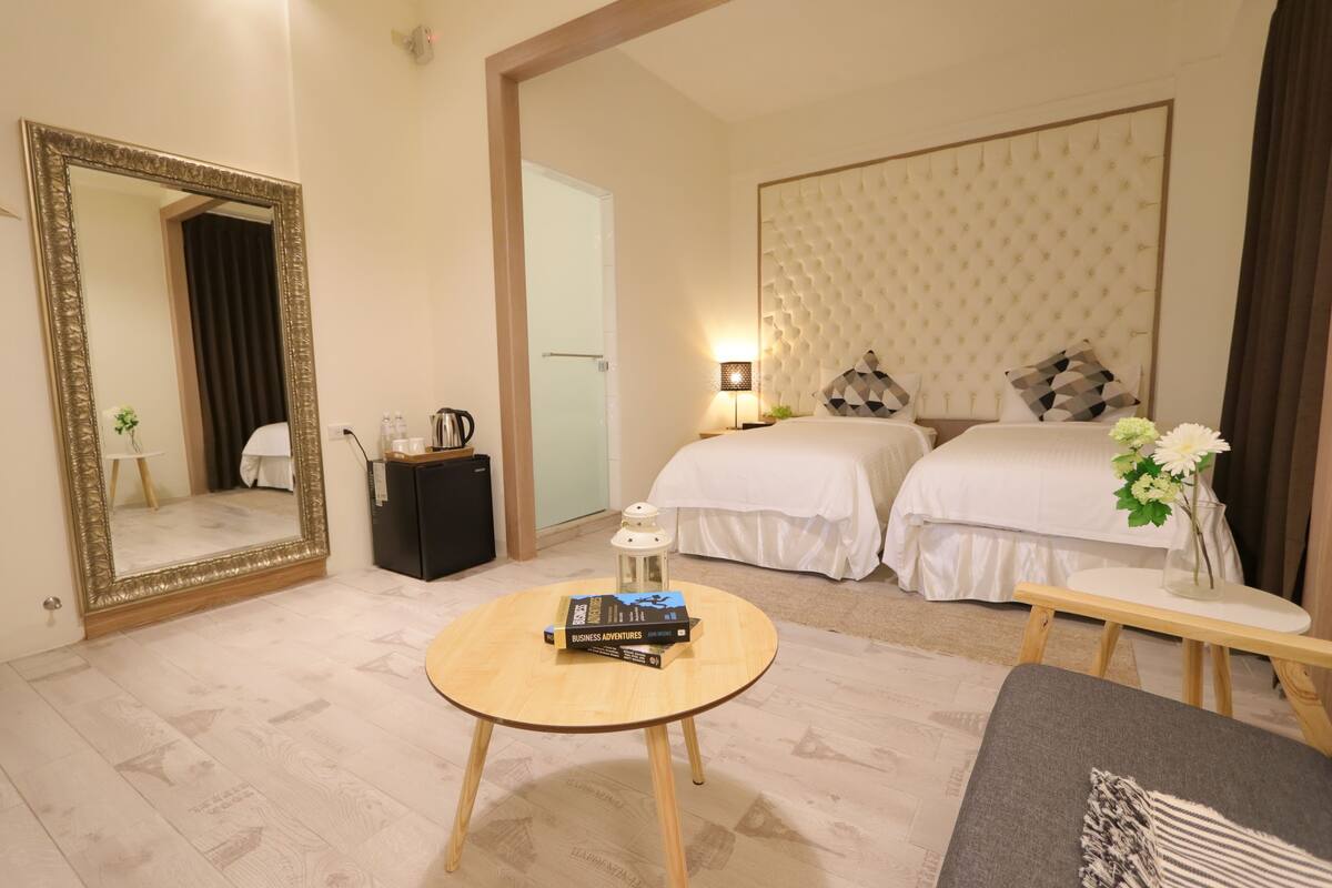 A hotel room with two beds, a round wooden table with a book, a mirror, and a bedside table with a lamp.