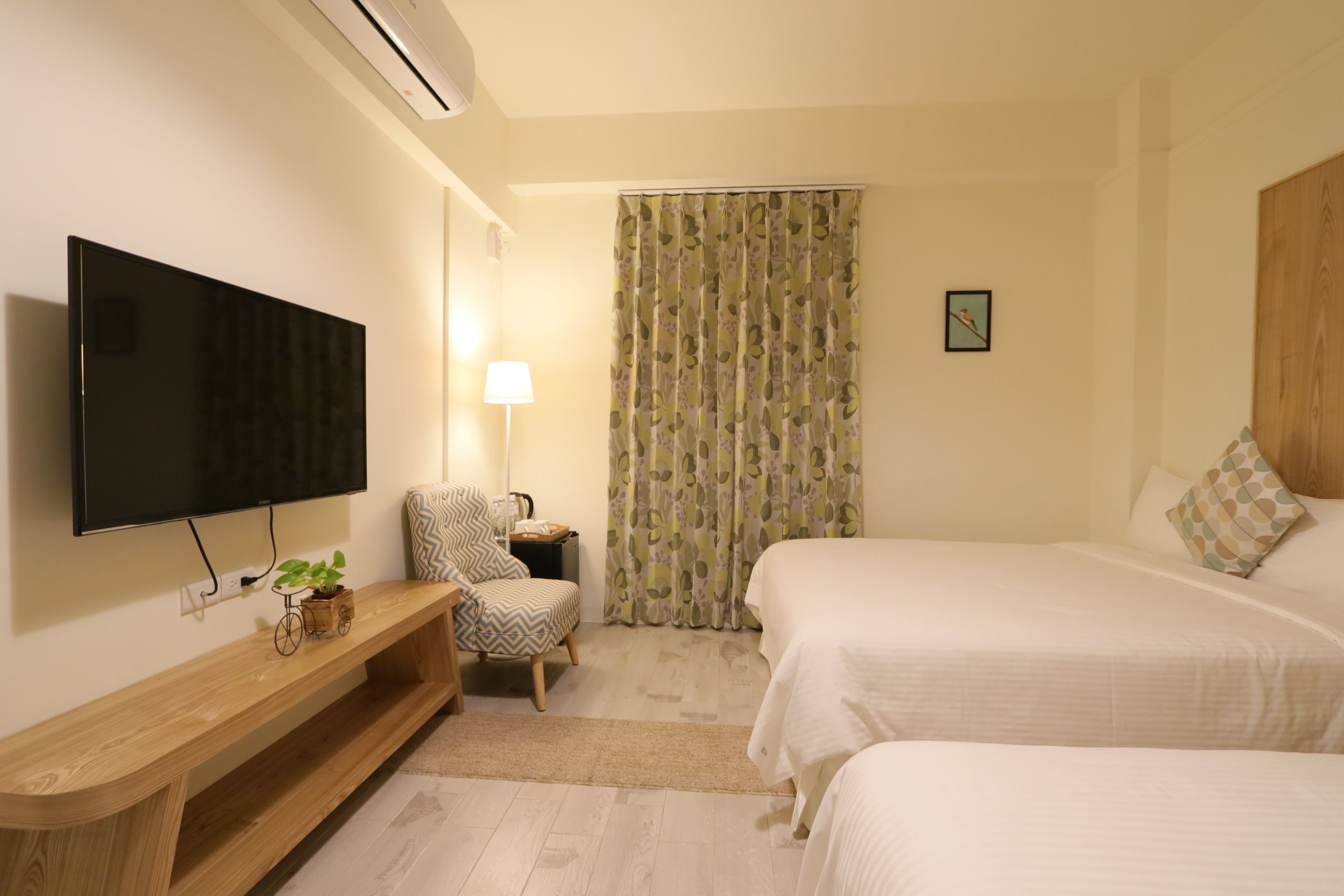 A hotel room with a flat-screen TV, a wooden console table with a plant, a patterned chair, a bedside table with a lamp, a bed with white bedding, and a curtain with a leaf pattern.