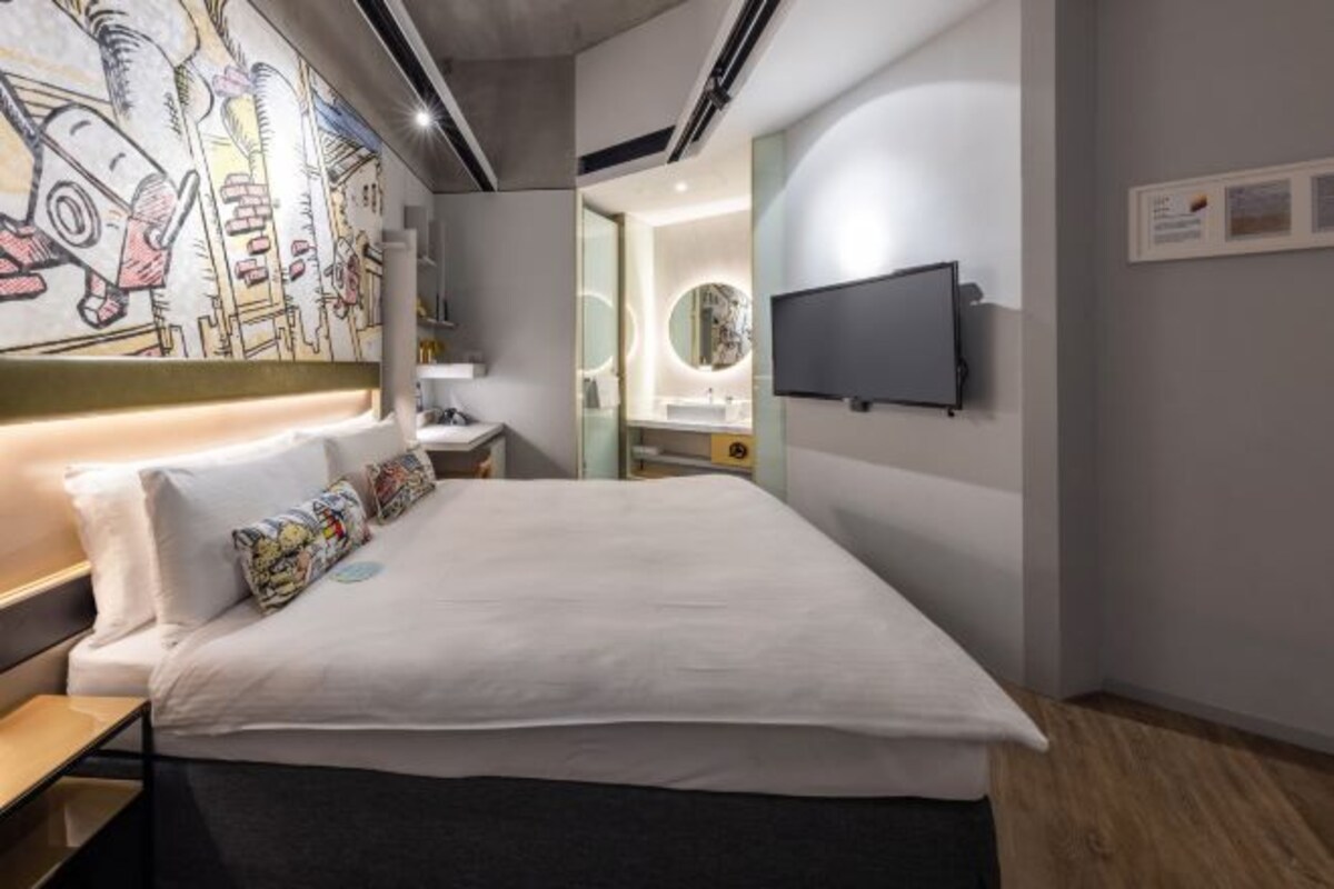A modern hotel room with a large bed, a TV mounted on the wall, and a unique mural on the wall.