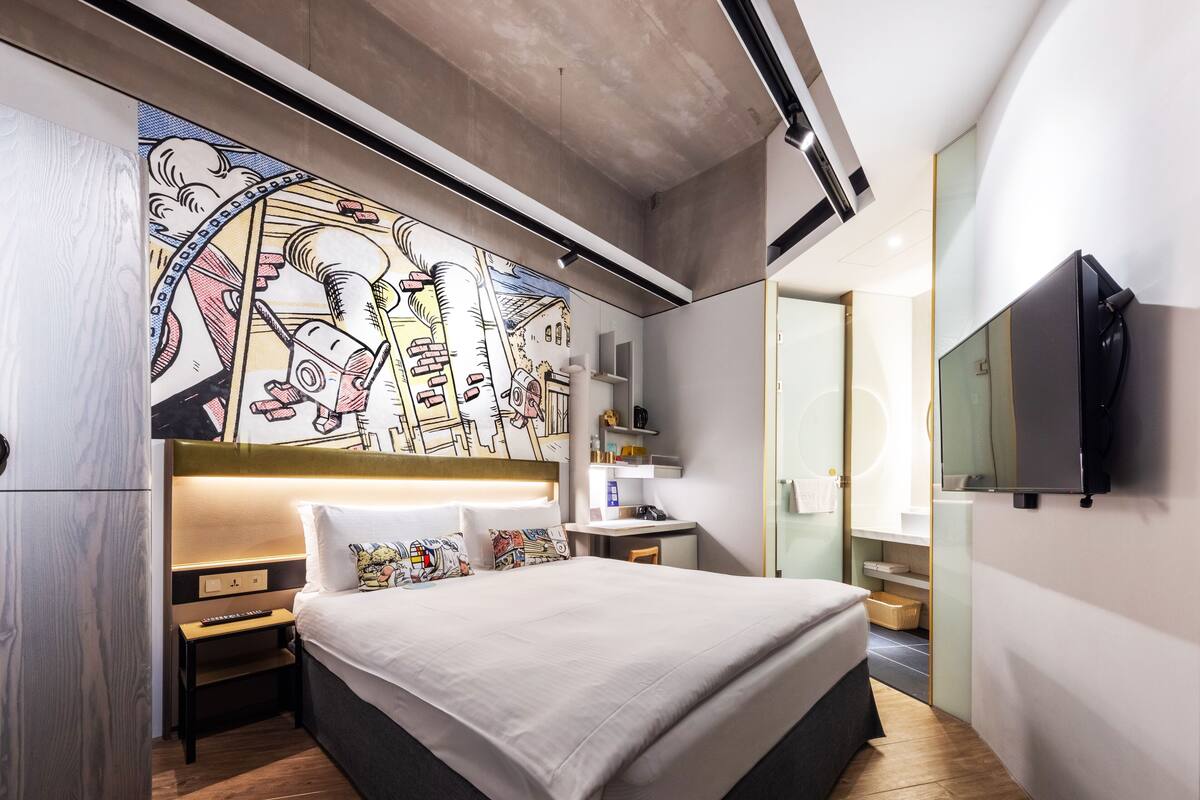 A modern hotel room with a large bed, a headboard with a mural, a TV mounted on the wall, and a wooden floor.