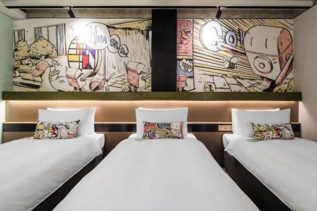 A hotel room with three beds, each with a colorful pillow. The room features a large mural on the wall depicting a comic-style scene.