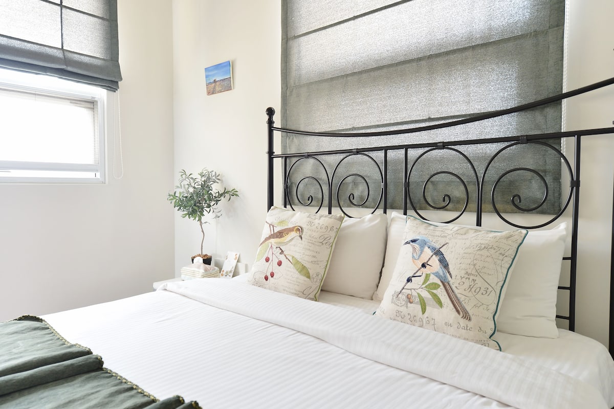 Una cama bien hecha con sábanas blancas y almohadones decorativos, una pequeña planta en la mesita de noche y una ventana con persianas.