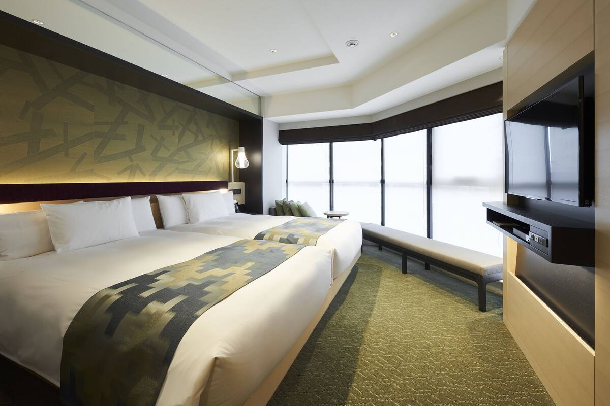 A hotel room with two beds, a large window, a flat-screen TV, and a bench.