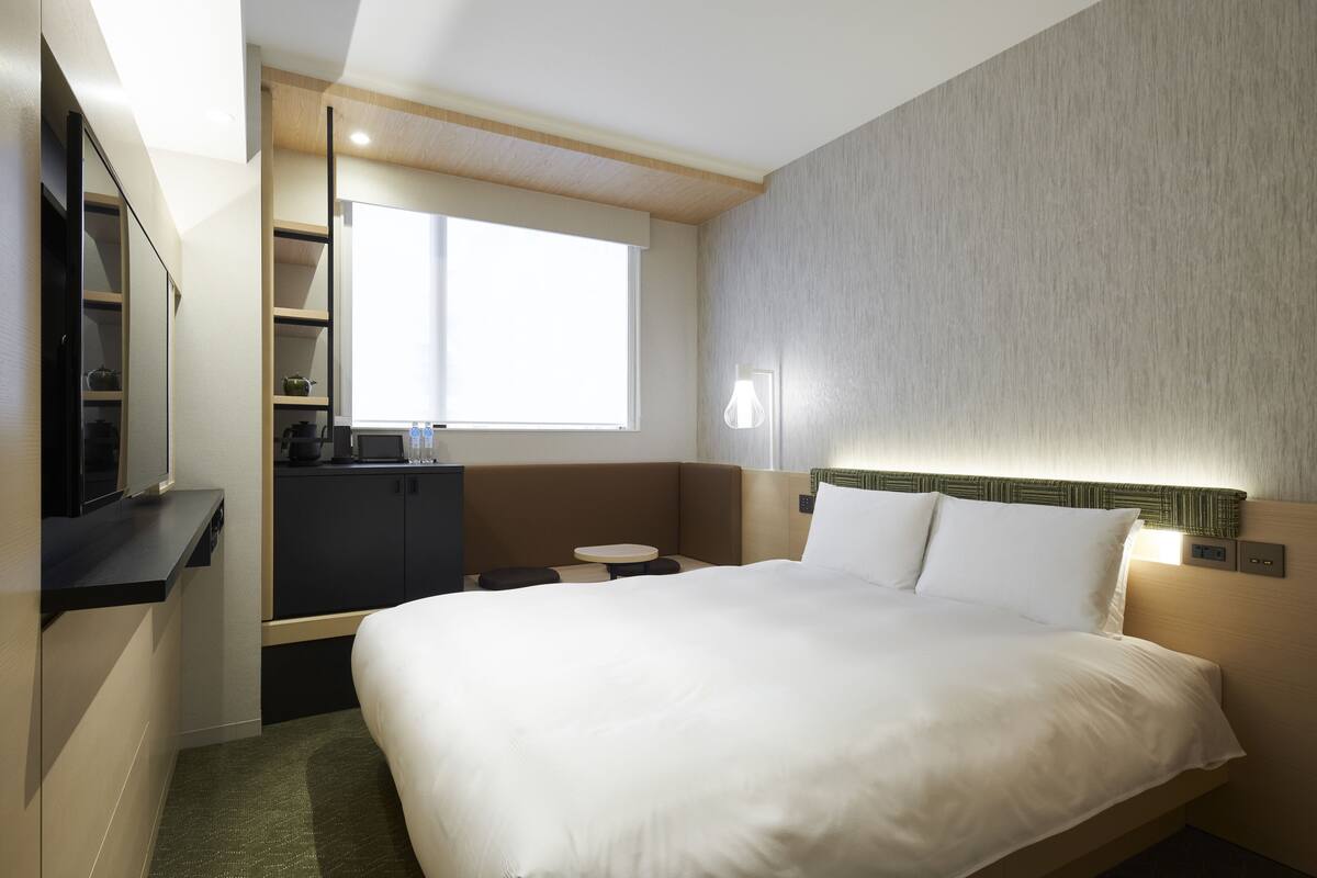 A hotel room with a large bed, a small table, a built-in TV, a kitchenette, and a window providing natural light.