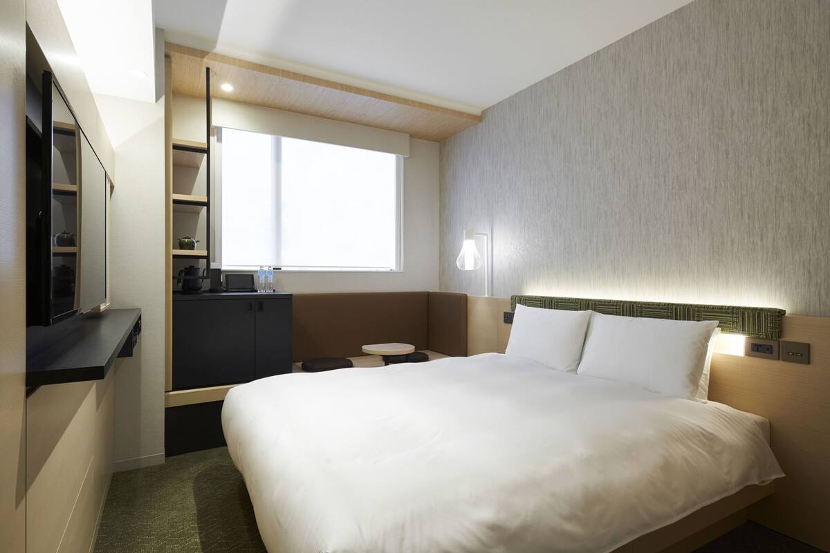 A hotel room with a bed, a desk, a small table, a TV, and a window.
