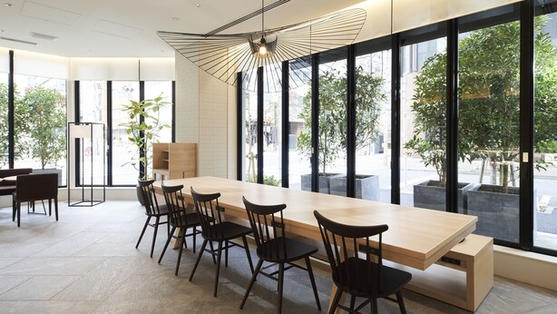 A modern dining area with a long wooden table, black chairs, and large windows offering a view of the street. - Hotel 1899 Tokyo (Tokyo)