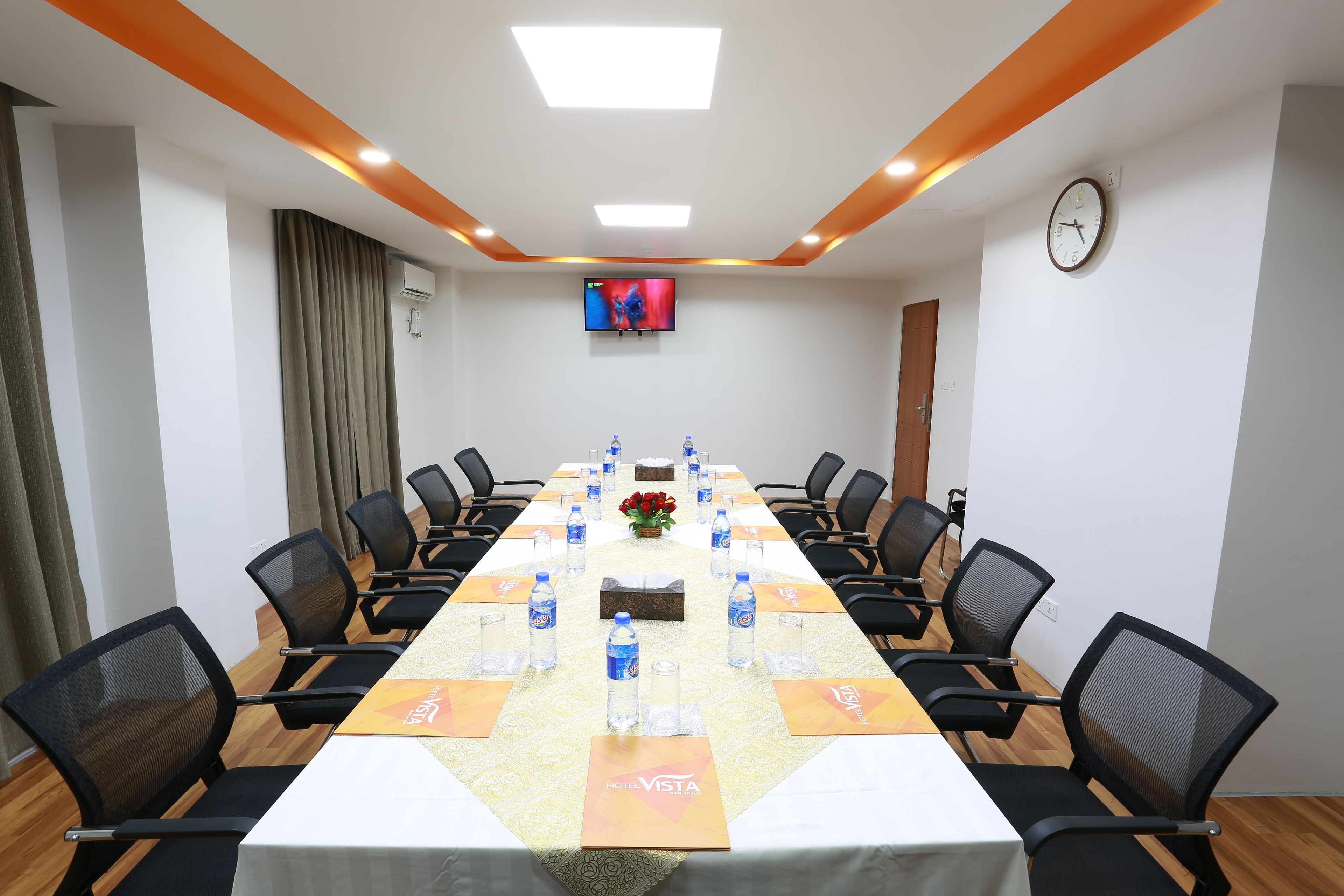 A conference room with a long table, black chairs, water bottles, a clock, and a TV on the wall.