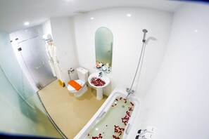 A modern bathroom with a glass shower enclosure, a bathtub with rose petals, a toilet with an orange seat, and a sink with a mirror above it. - Hotel Vista (Yangon)