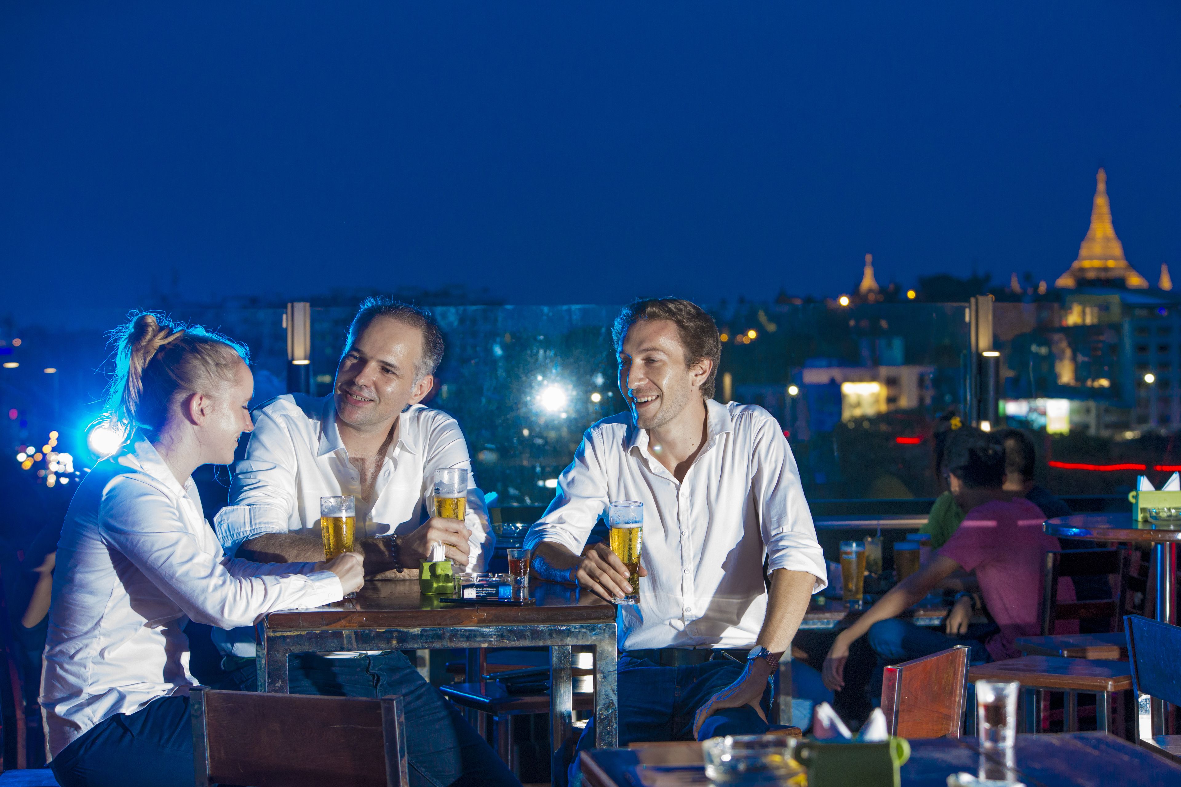 A group of people sitting at a table with drinks, in an outdoor setting with a cityscape in the background.