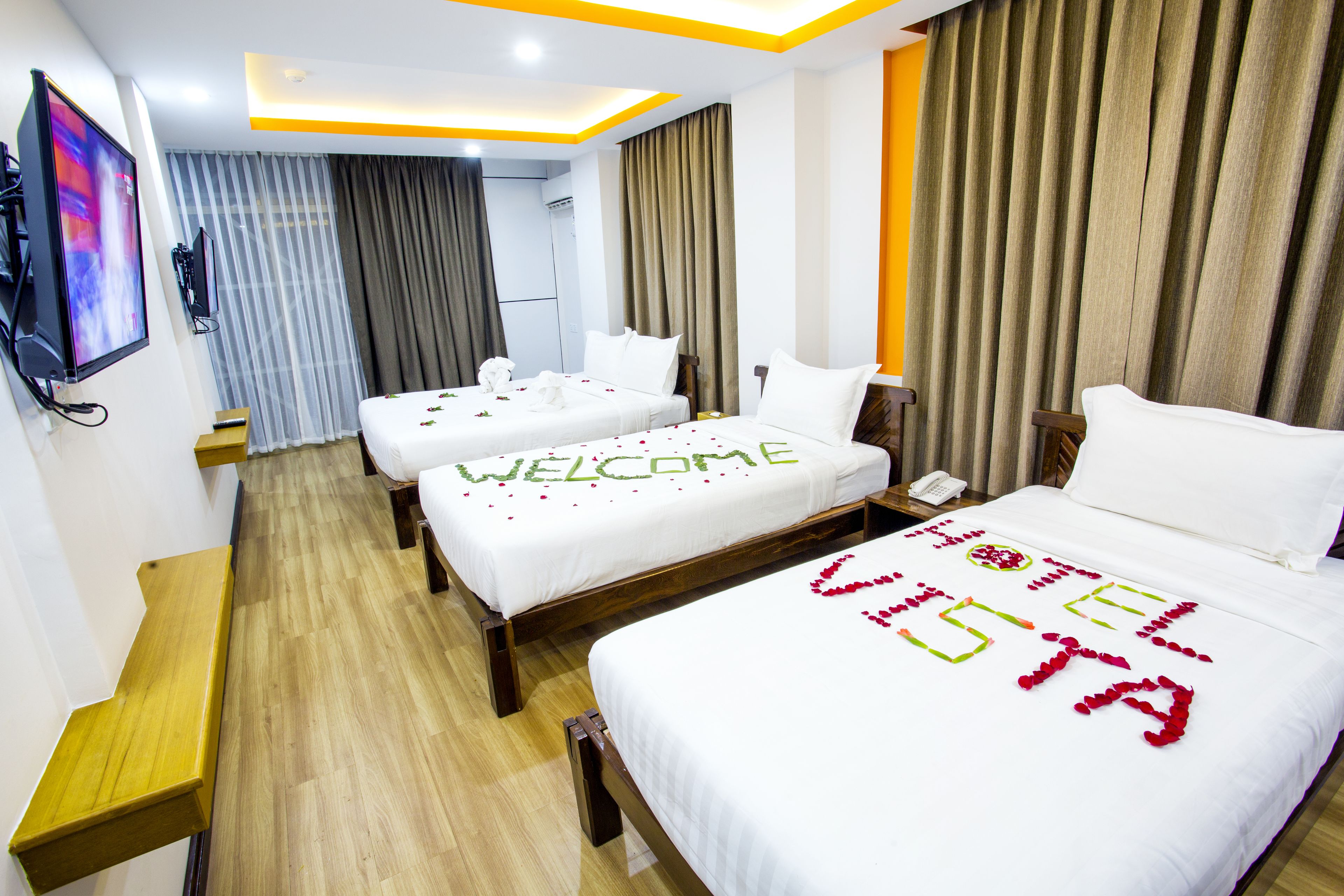 A hotel room with two beds, a TV, a wooden bench, and a "WELCOME" sign on the beds.