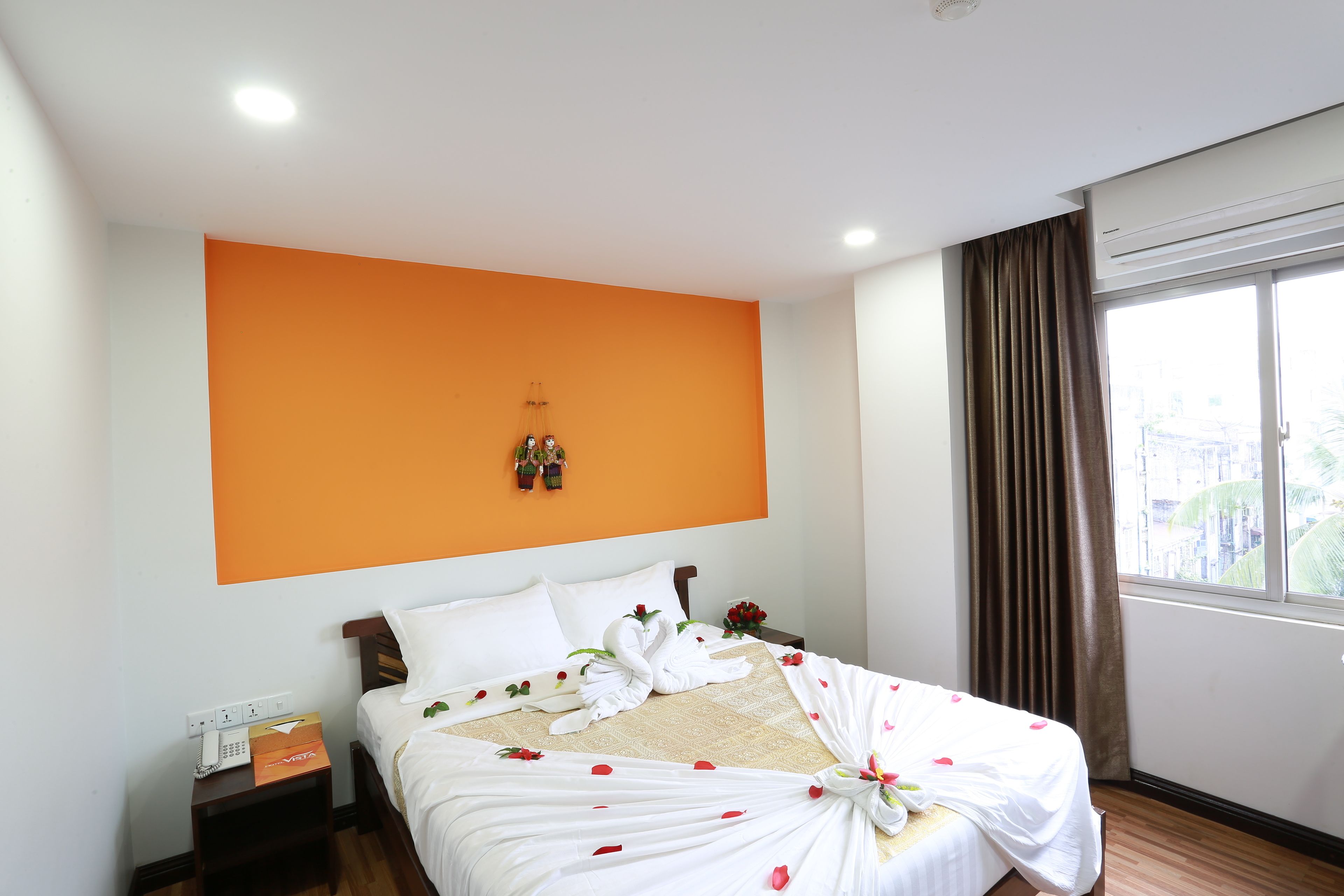 A hotel room with an orange accent wall, a neatly made bed with white linens and rose petals, a small table with a telephone, and a window with curtains.