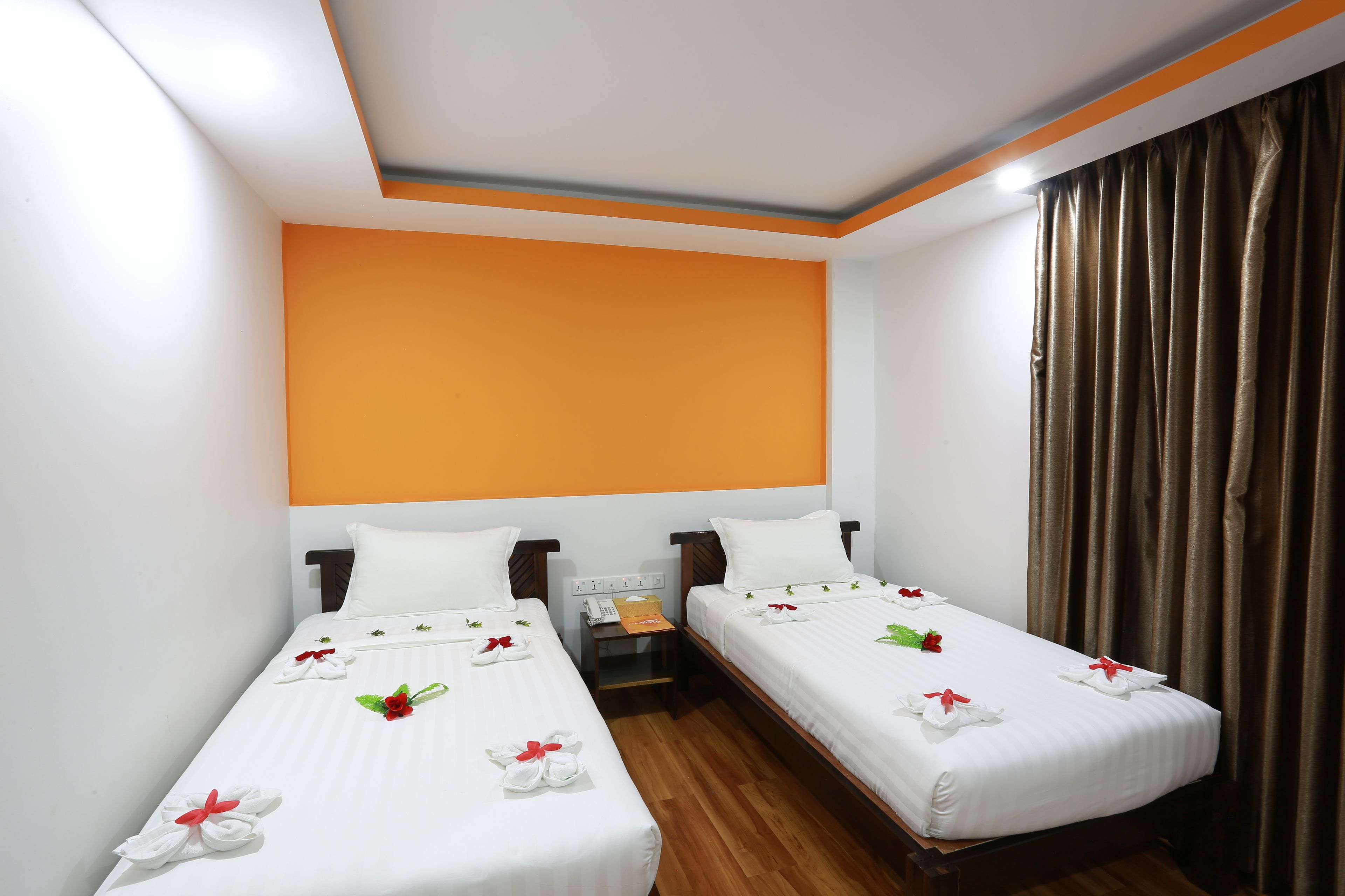 A hotel room with two single beds, each with white linens and red bows. There is a wooden nightstand between the beds with a phone and a small box. The room has an orange accent wall and a brown curtain.