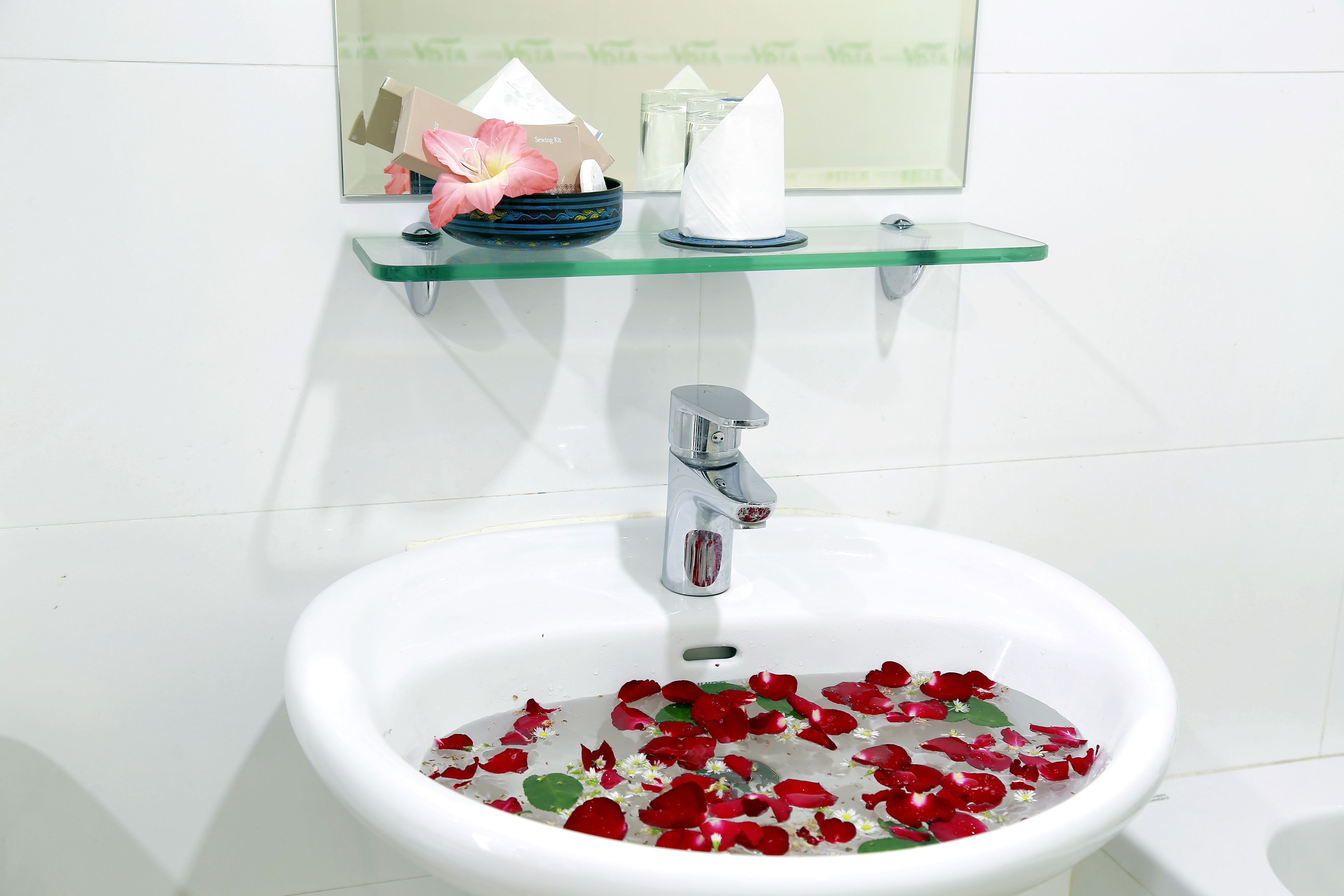 A bathroom sink with water and rose petals, a mirror, and a glass shelf with a flower and a tissue box.