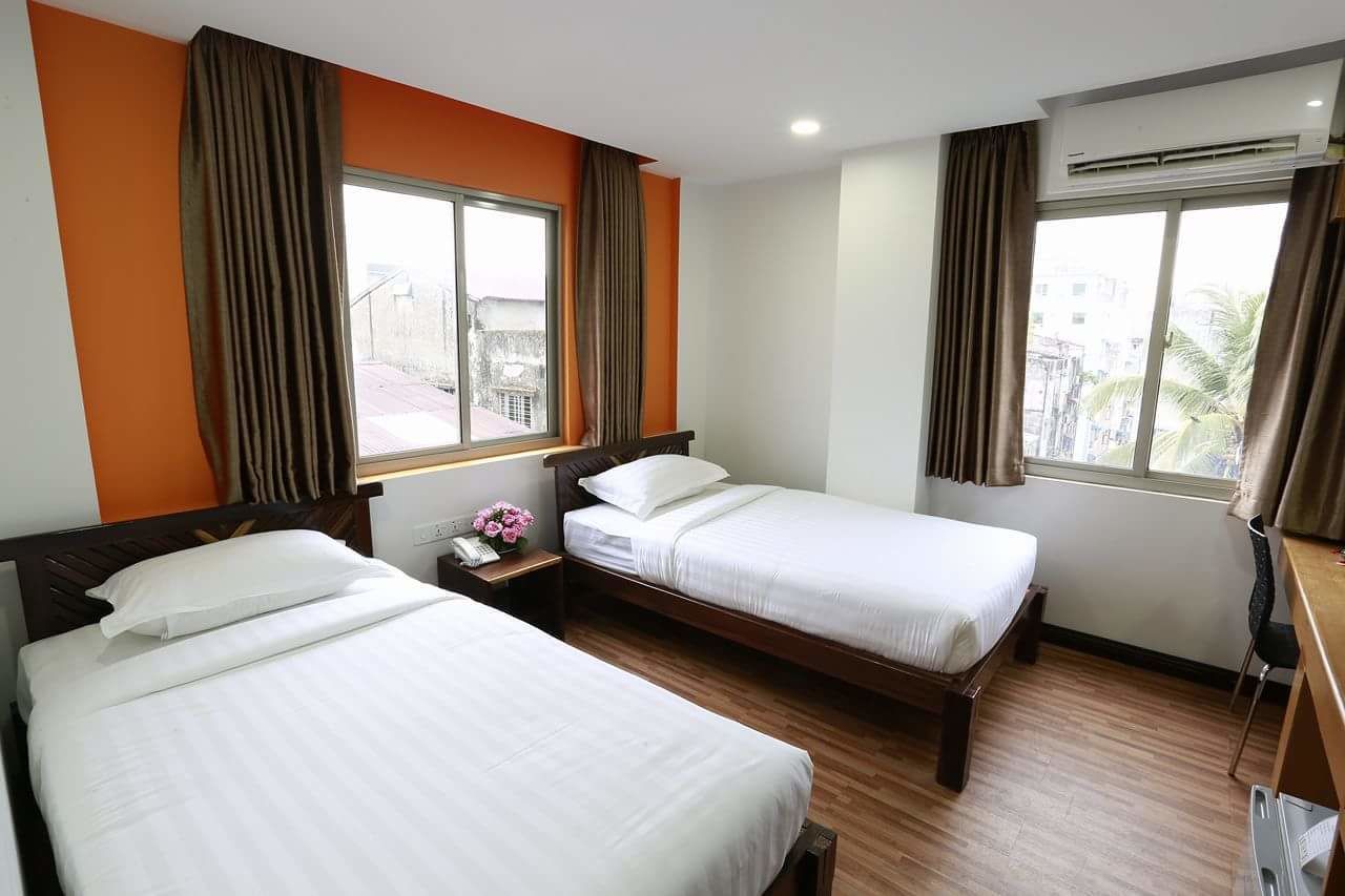 A hotel room with two single beds, a wooden floor, a small bedside table with a lamp, and a window with curtains.