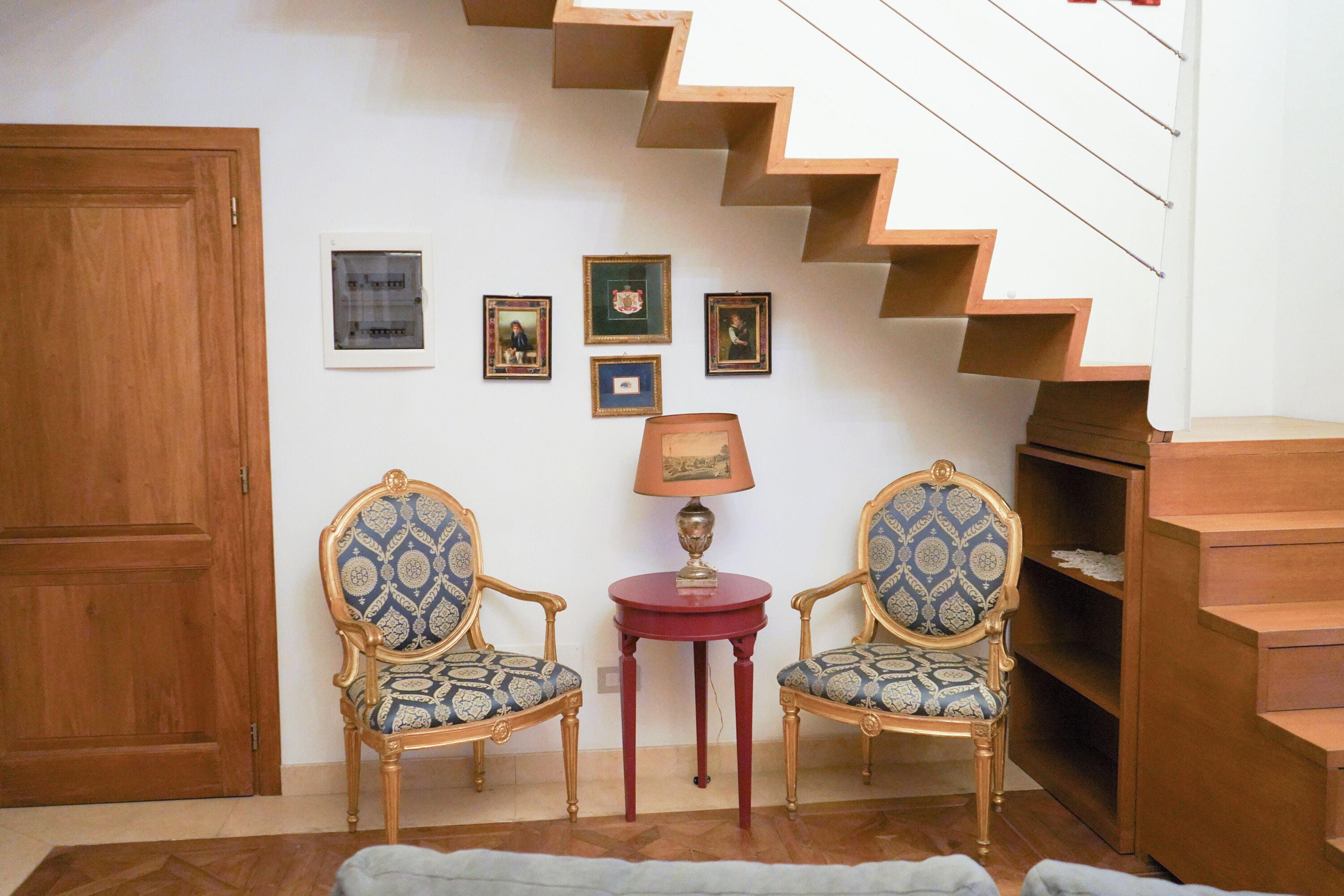 A room with a wooden staircase, two ornate chairs, a small table with a lamp, and framed pictures on the wall.