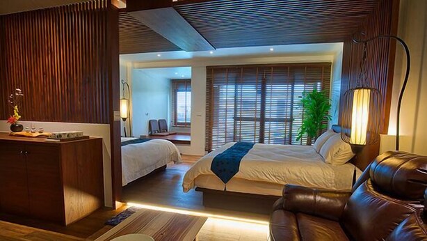 Quarto de hotel moderno com escrivaninha de madeira, duas camas, sofá de couro e uma grande janela com persianas.