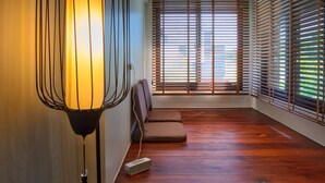 Quarto moderno com piso de madeira, um banco, luminária fixada na parede e amplas janelas com persianas.