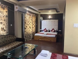 A hotel room with a sofa, a glass coffee table, a bed with a headboard, and curtains. - Hotel Shefaf (Srinagar)