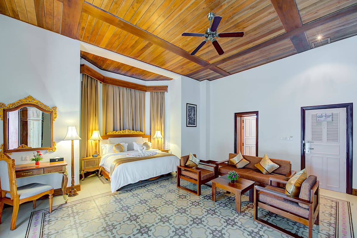 A hotel room with a bed, two sofas, a desk, a chair, a ceiling fan, and a patterned floor.