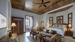Habitación de hotel con techo de madera, cama grande, escritorio con lámpara, una silla y un sofá. Hay un cuadro enmarcado en la pared y una ventana con persianas.