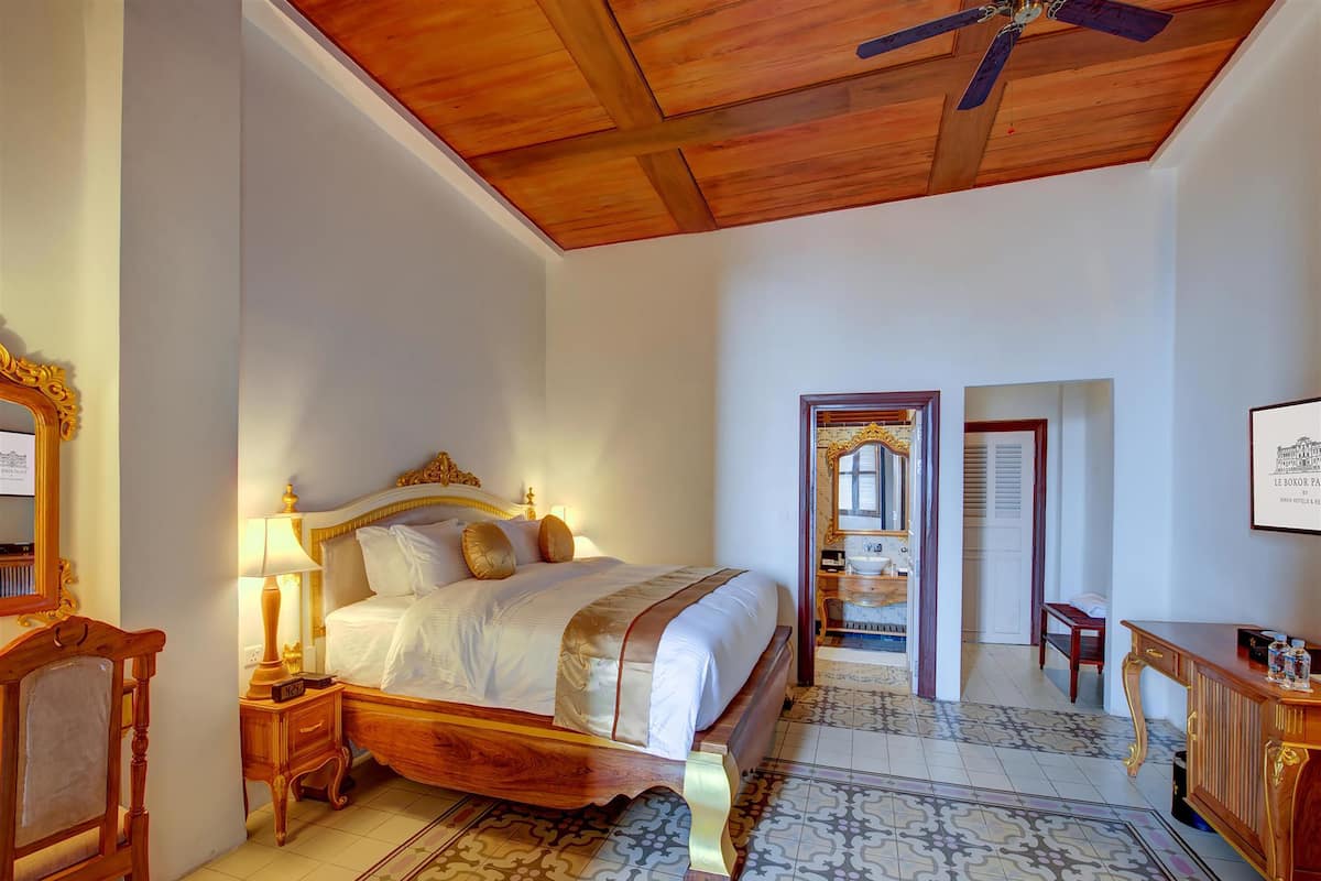 A bedroom with a wooden bed, a nightstand, a chair, and a desk. There is a ceiling fan and a door leading to another room.